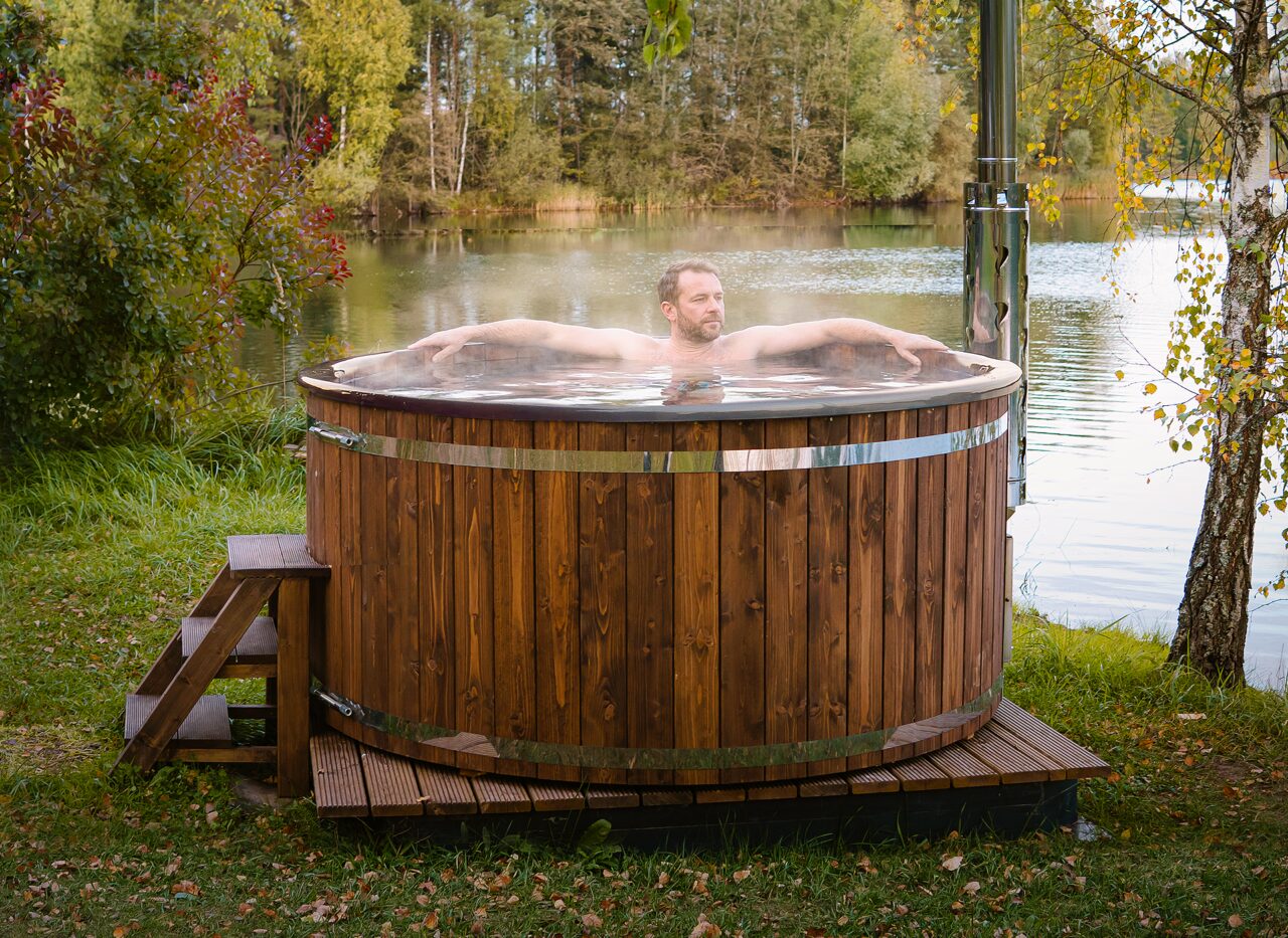 Eine Person entspannt in einem holzbefeuerten Feuerbad Hot Tub SOPHIA auf Holz am Ufer eines Sees, mit Metallschornstein, Holz-Einstiegstreppe und Wald im Hintergrund