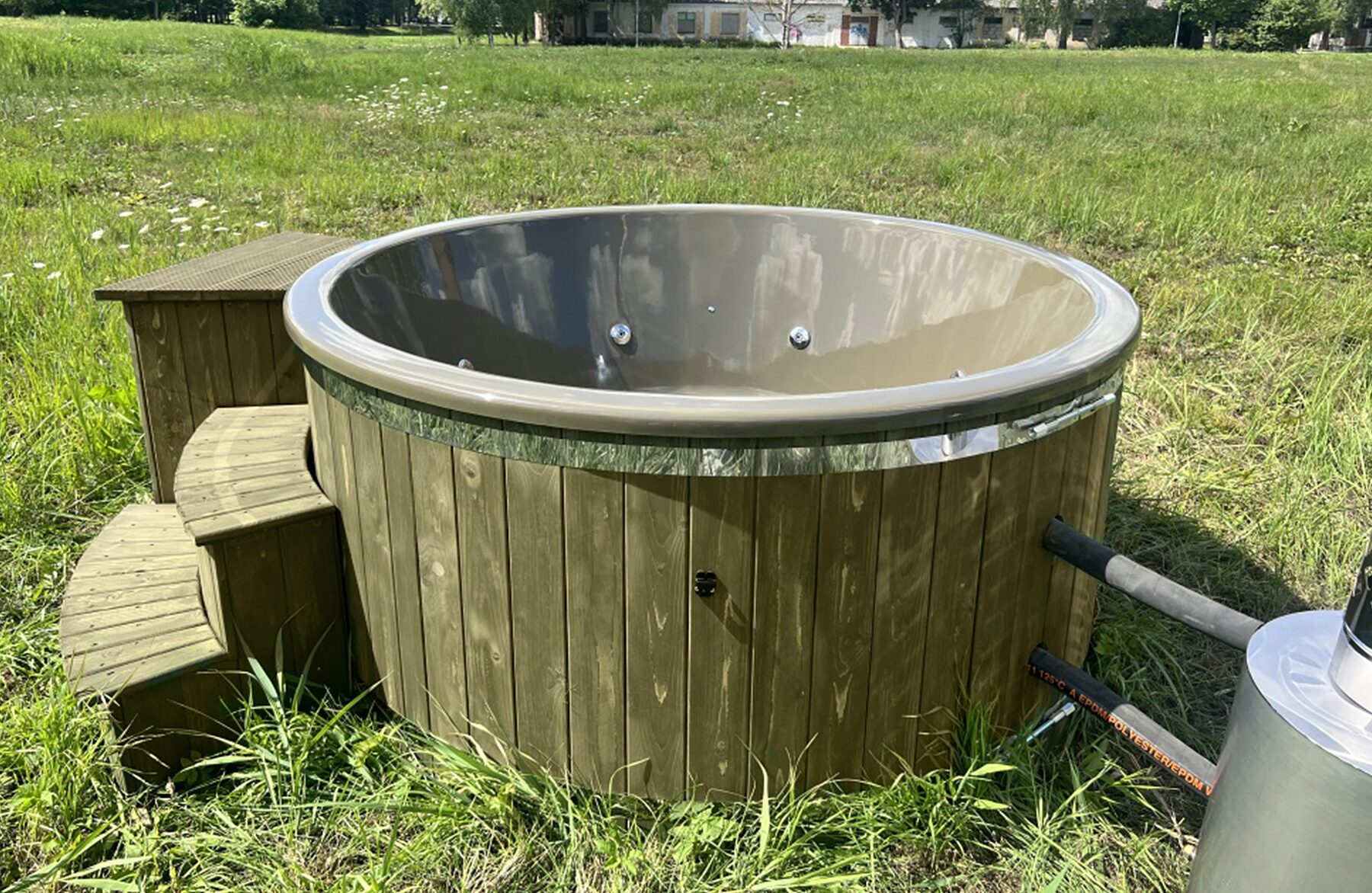 Feuerbad Hot Tub DIANA auf einer grünen Wiese, mit Holzverkleidung, grauer Innenwanne mit integrierten Düsen, Holz-Einstiegstreppe und externem Metallofen