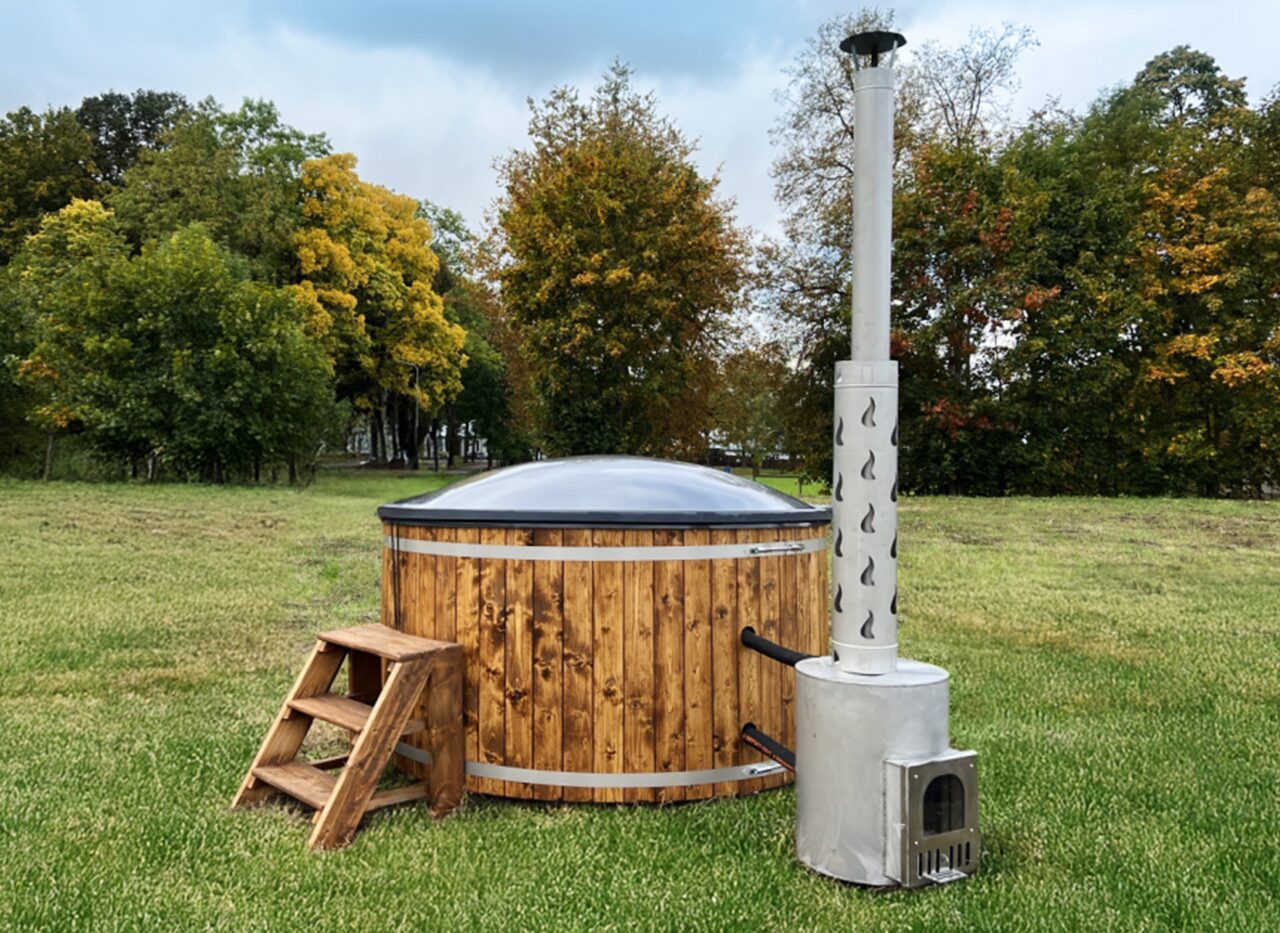 Feuerbad Hot Tub DIANA steht auf einer grünen Wiese und verfügt über eine Holzverkleidung, eine Holz-Einstiegstreppe, eine isolierende Abdeckung sowie einen externen Metallofen mit hohem Schornstein, umgeben von einer natürlichen Landschaft