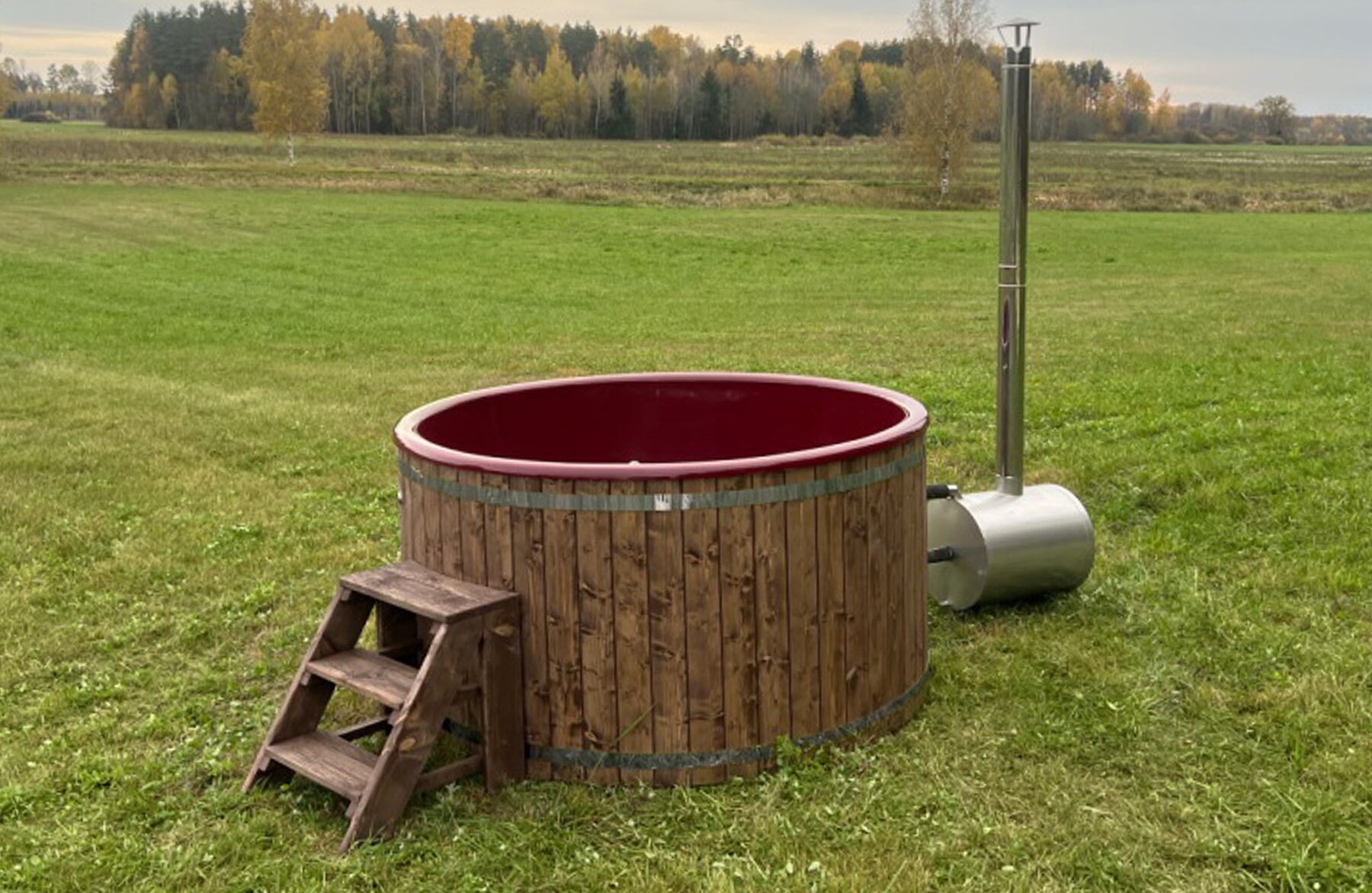 Feuerbad Hot Tub LUNA aus Holz mit roter Innenwanne auf einer Wiese, mit externem Metallofen und Schornstein, Holz-Einstiegstreppe und grüner Naturlandschaft im Hintergrund
