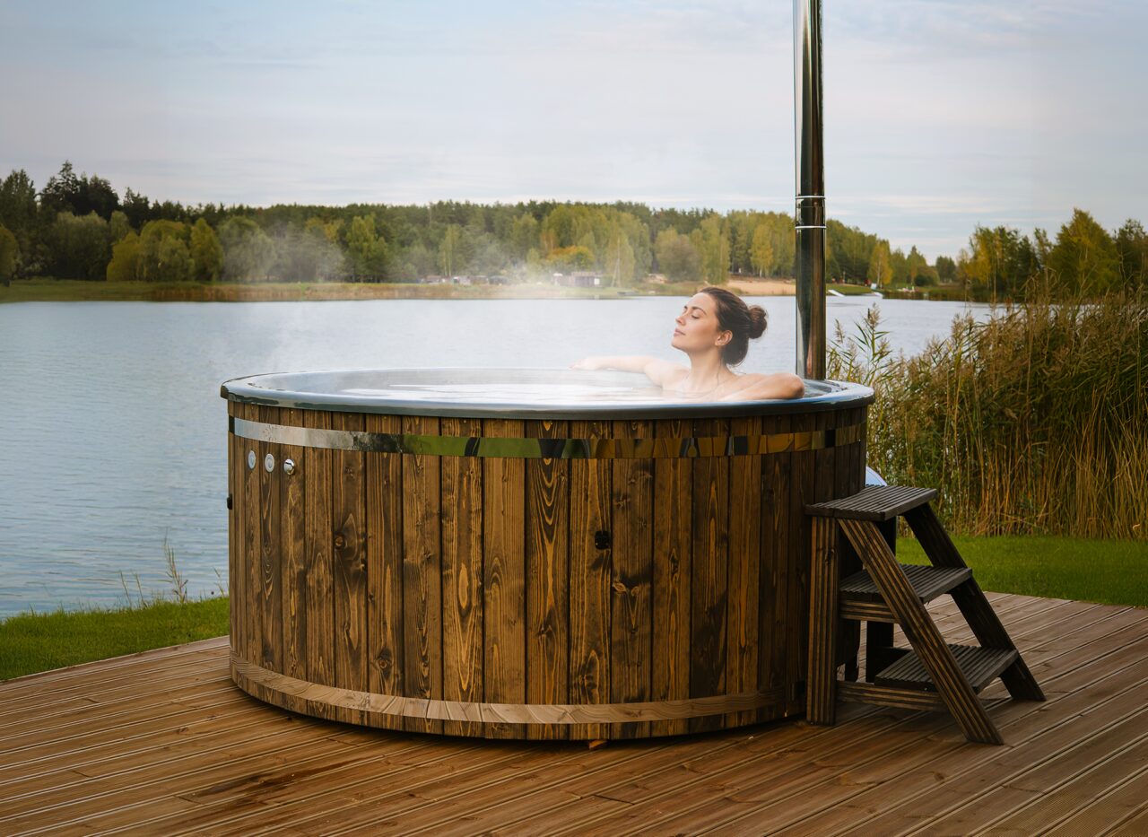 Eine Person entspannt im warmen Wasser des Feuerbad Hot Tubs LUNA auf einer Terrasse am See, mit Holzverkleidung, Metallschornstein, Einstiegstreppe und ruhiger Naturlandschaft im Hintergrund