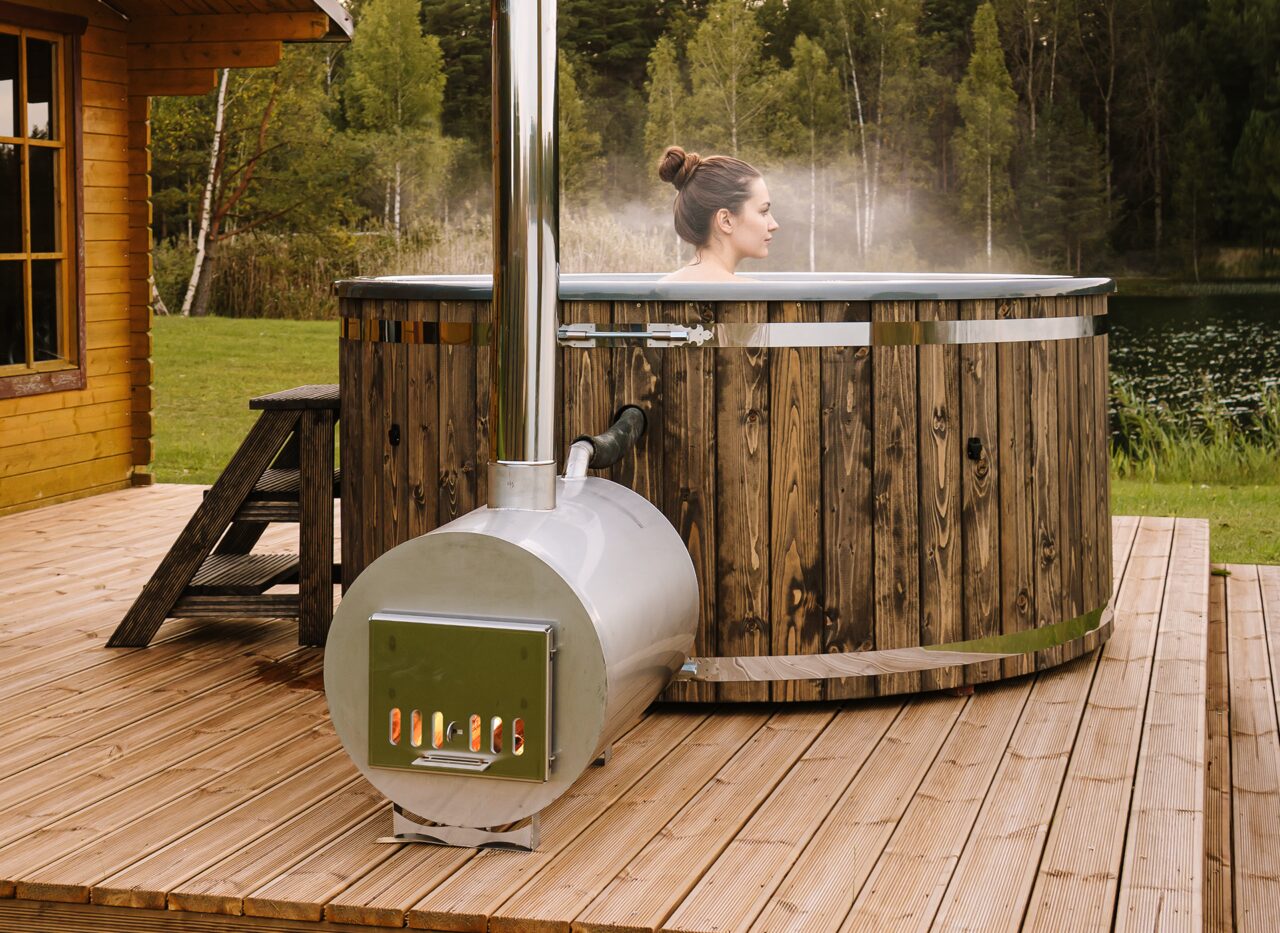 Eine Person entspannt im Feuerbad Hot Tub LUNA auf einer Terrasse neben einem Holzhaus, mit Holzverkleidung, externem Metallofen mit Schornstein, Einstiegstreppe und Landschaft im Hintergrund