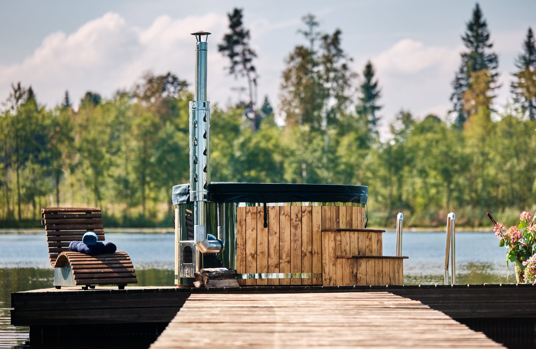 Feuerbad Hot Tub SOPHIA mit schwarzer Abdeckung auf einem Holzsteg am See, mit Metallofen und Schornstein, Einstiegstreppe und Liegestuhl, umgeben von Wald