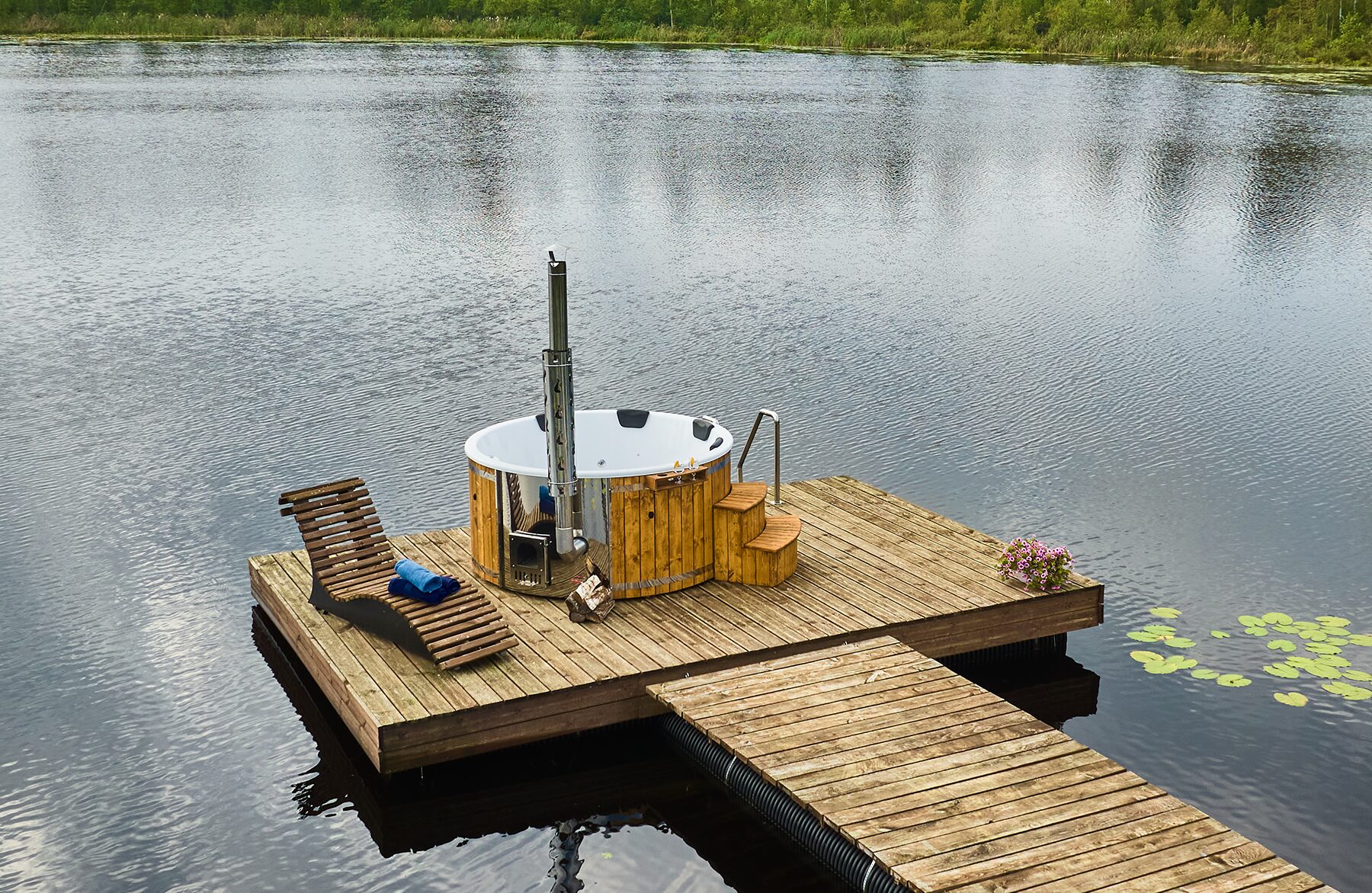 Feuerbad Hot Tub SOPHIA mit weißer Innenwanne auf einem Steg am See, mit Metallofen und Schornstein, Einstiegstreppe, Liegestuhl und Handtüchern, umgeben von Natur