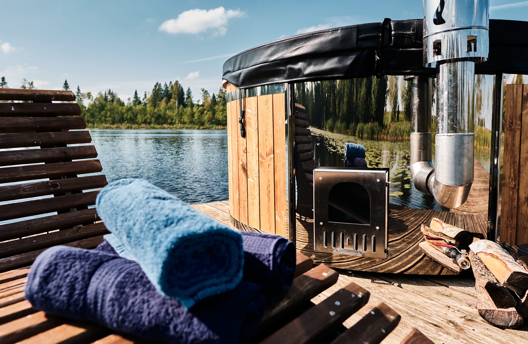 Detailansicht Feuerbad Hot Tub SOPHIA auf einem Holzsteg am See, mit schwarzer Abdeckung, Metallofen und Schornstein, Brennholz sowie zusammengelegten Handtüchern auf einer Holzliege