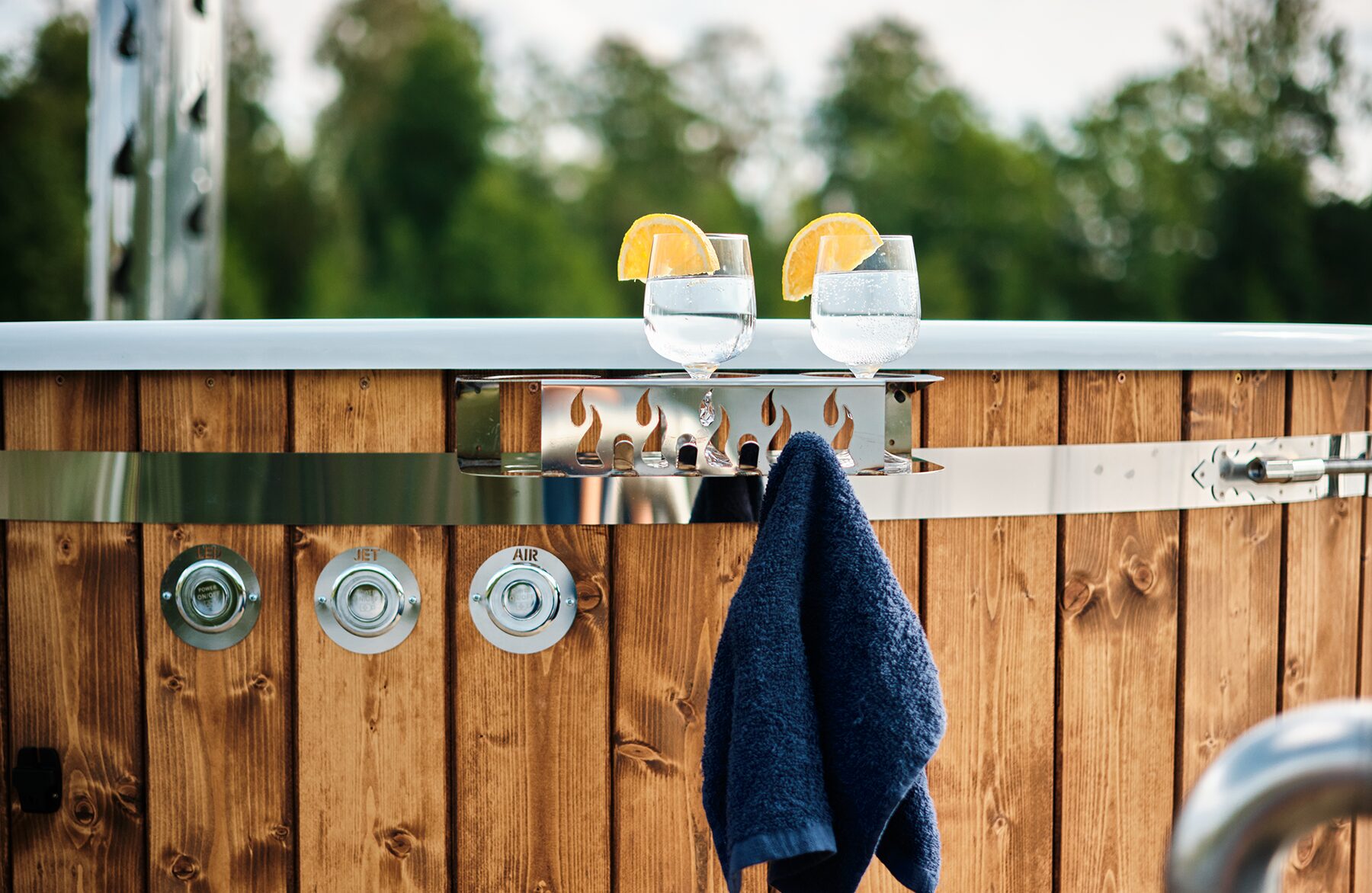 Detailansicht Feuerbad Hot Tub SOPHIA mit Getränkeablage, zwei Gläsern Wasser mit Orangenscheiben, Handtuchhalter und Holzverkleidung vor grünem Hintergrund