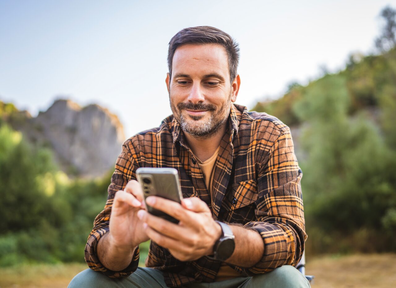 Eine Person sitzt im Freien und schaut lächelnd auf ein Smartphone