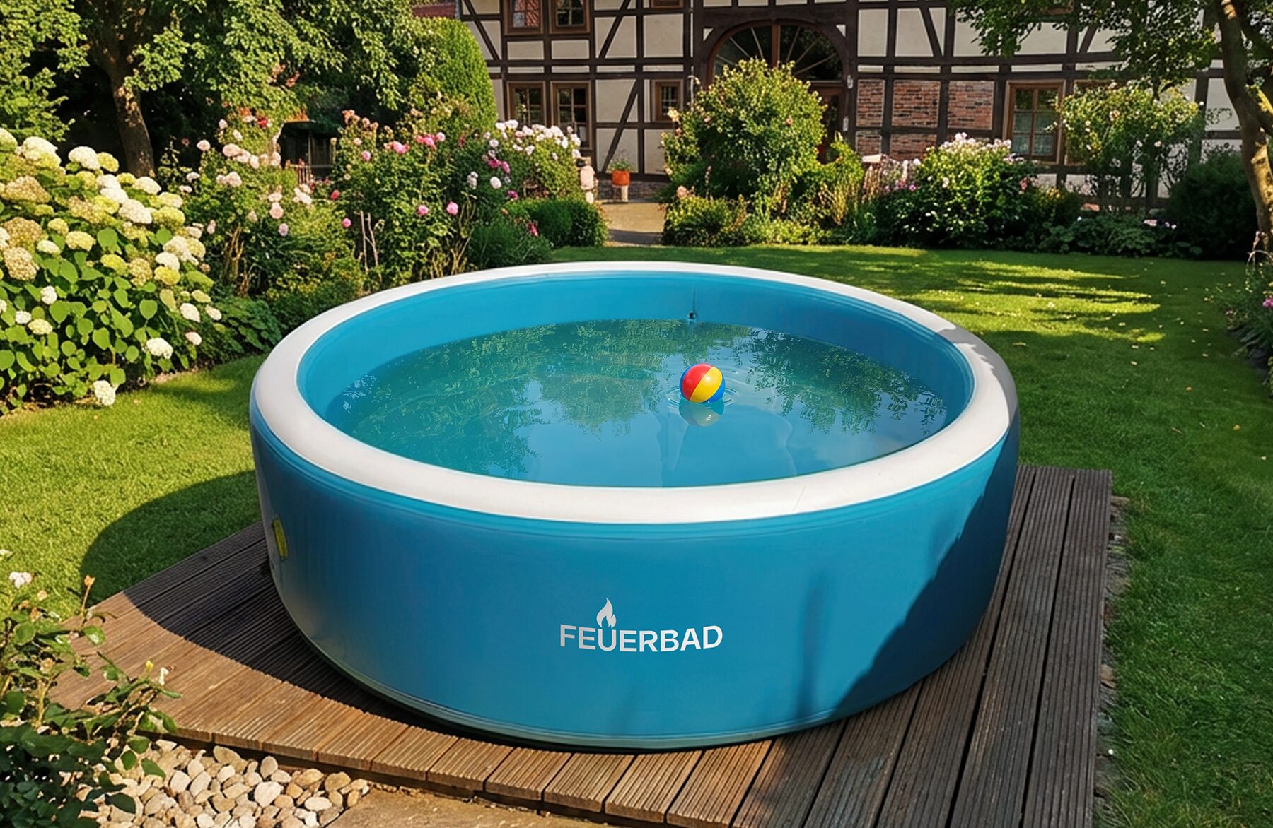 Außenansicht eines runden, aufblasbaren Pools von Feuerbad mit blauer Außenwand und klarem Wasser, platziert auf einer Holzterrasse in einem gepflegten Garten mit Rasen, Blumenbeeten und einem Fachwerkhaus im Hintergrund.