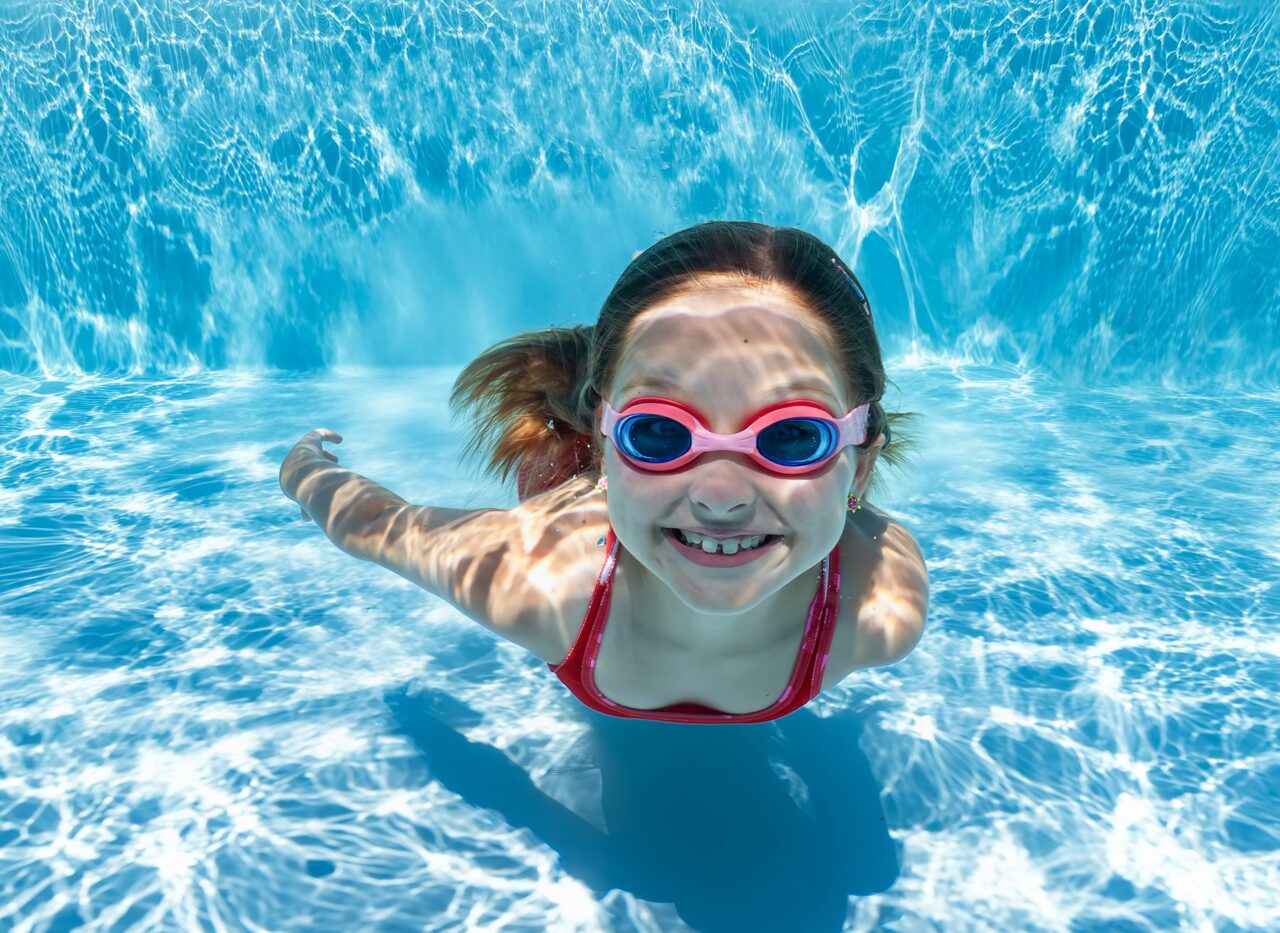 Ein Kind taucht lächelnd unter Wasser in einem klaren, blauen Pool und trägt eine Schwimmbrille, während Lichtreflexe auf der Wasseroberfläche und am Poolboden spielen.
