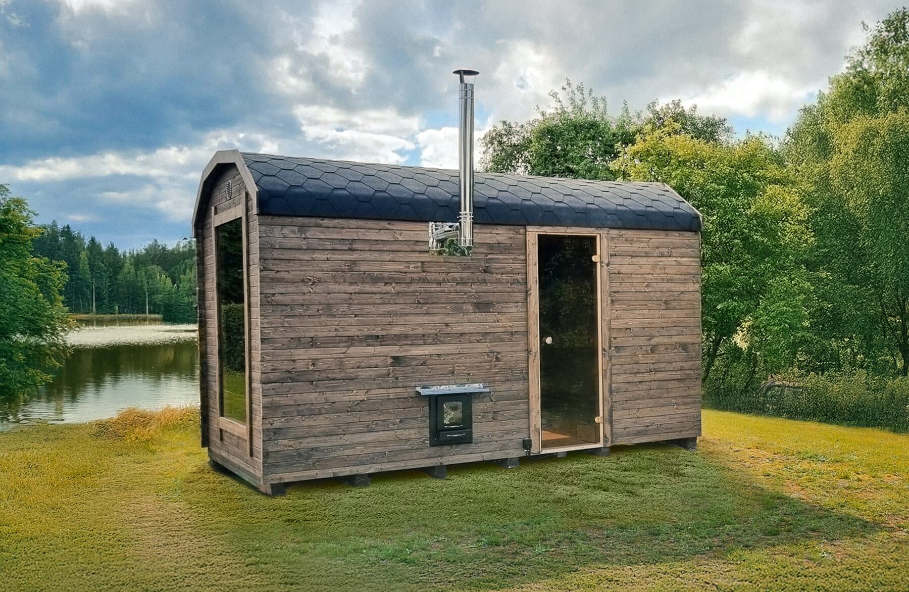 Feuerbad Sauna CERES auf einer Wiese, umgeben von Büschen und Bäumen. Im Hintergrund sieht man einen See und blauen Himmel