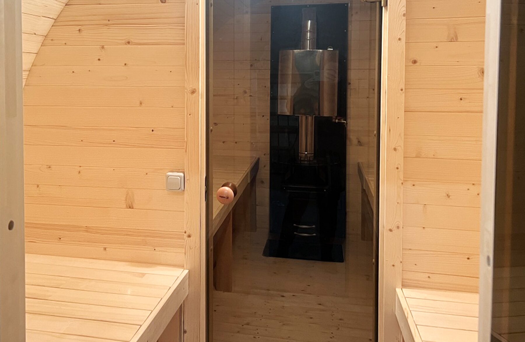 Innenansicht der Feuerbad Fass-Sauna JUNO mit heller Holzverkleidung, gegenüberliegenden Sitzbänken und Blick durch die Glastür auf den integrierten Saunaofen im Vorraum