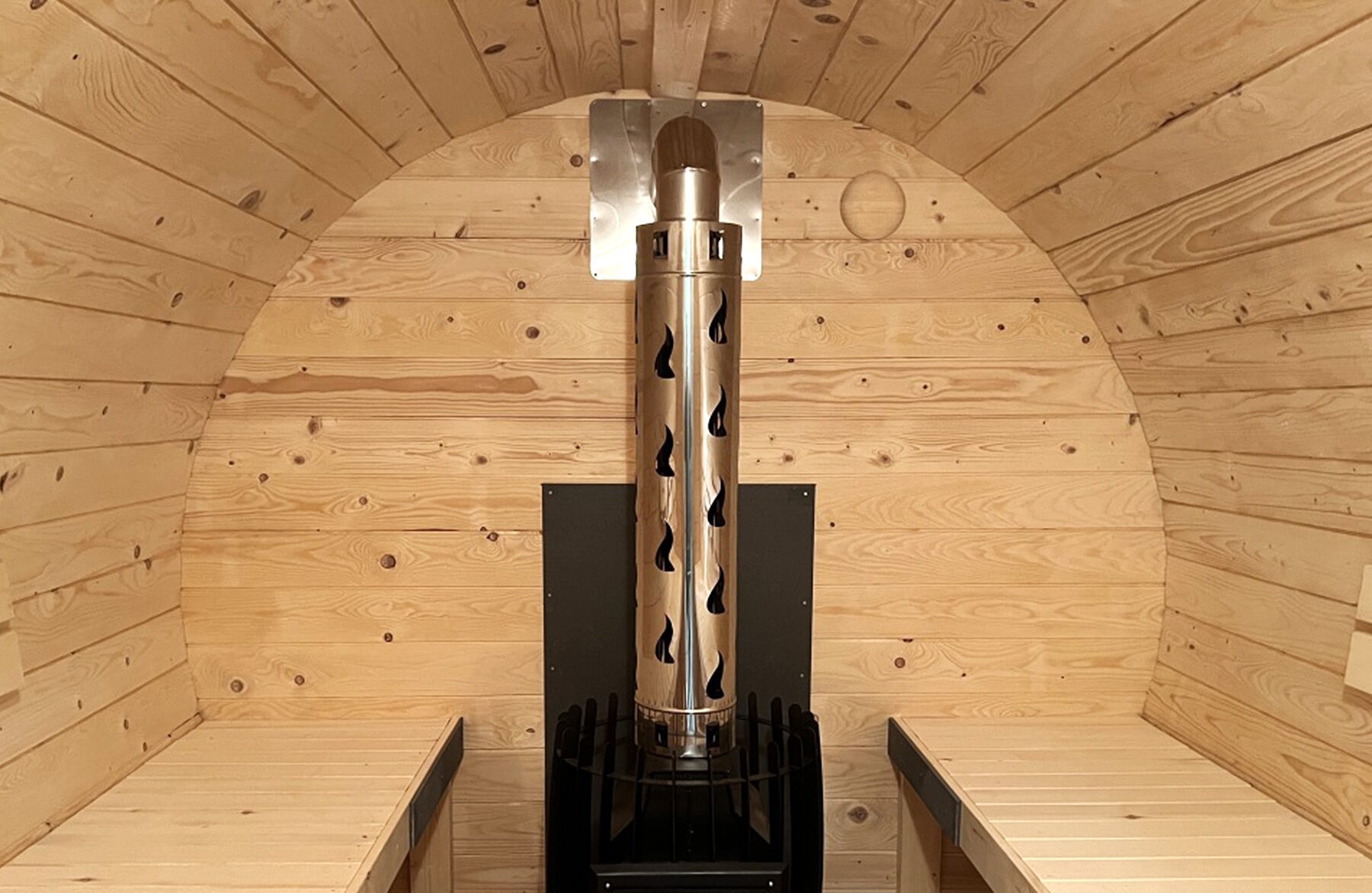 Zentrale Innenansicht der Feuerbad Fass-Sauna JUNO mit integriertem Holzofen und Schornstein, umgeben von heller Holzverkleidung und gegenüberliegenden Sitzbänken