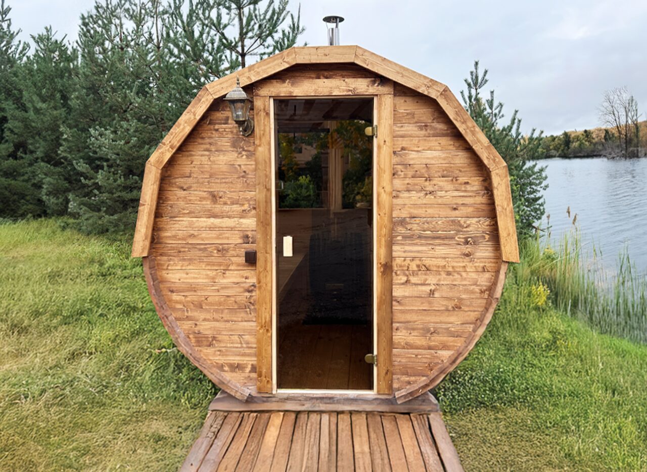 Frontansicht der Feuerbad Sauna JUNO mit runder Holzform, Glastür, dunkler Dachschindel und Holzdeck, auf einer grünen Wiese direkt am See, umgeben von Bäumen und ruhiger Natur