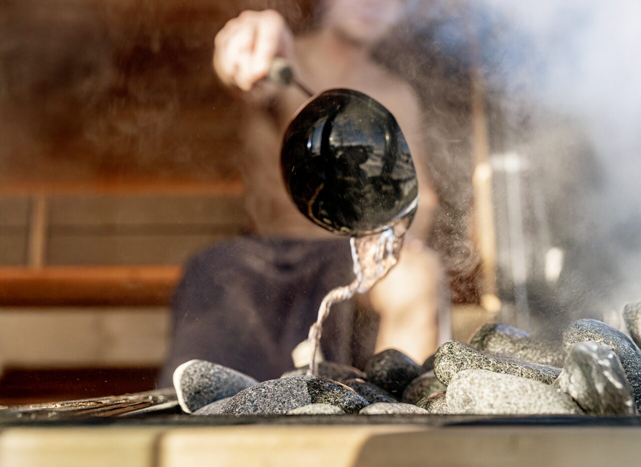 Eine Person gießt Wasser mit einer Kelle über heiße Saunasteine im Inneren der Feuerbad Sauna JUNO, wodurch aufsteigender Dampf entsteht
