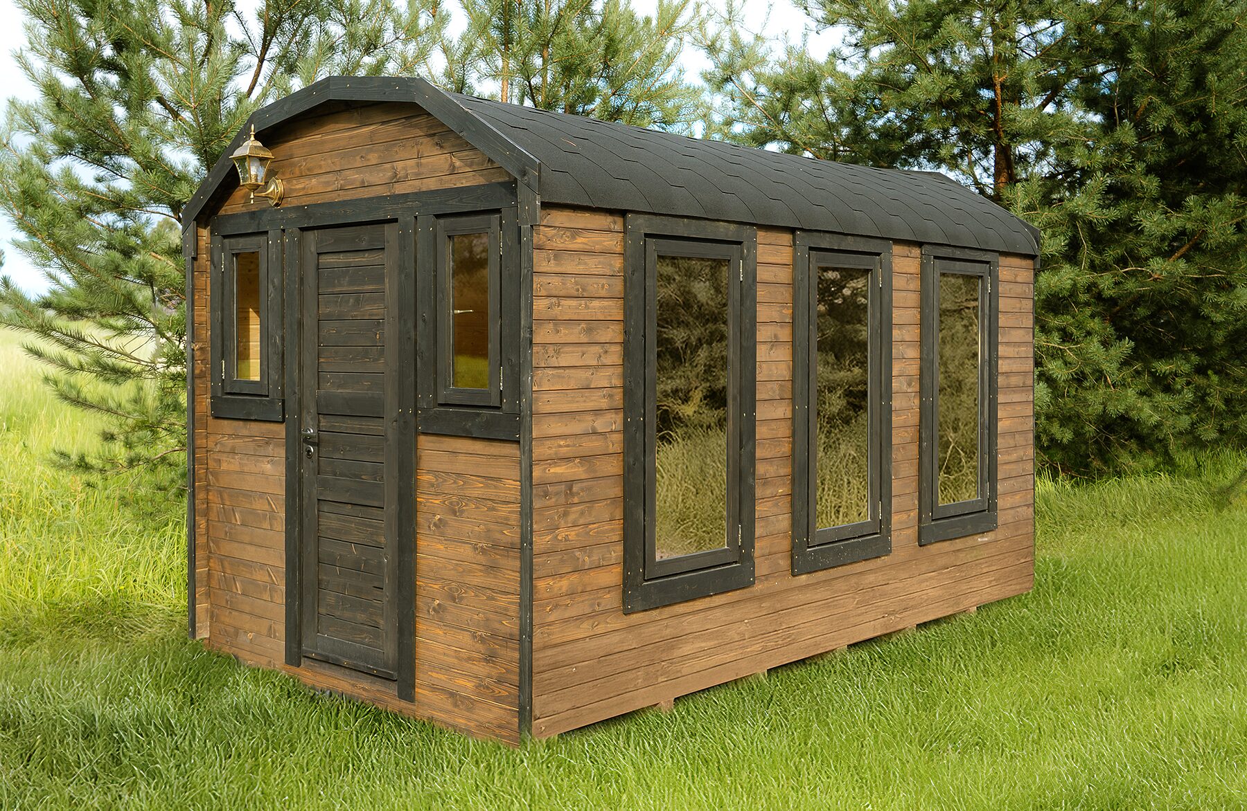Außenansicht eines Feuerbad Tiny House CERES mit dunklem Holzrahmen und warmer Holzverkleidung. Das kompakte Haus steht auf einer grünen Wiese, umgeben von Kiefern. Große Fenster sorgen für viel Tageslicht, das dunkle, leicht geneigte Dach unterstreicht den modernen, natürlichen Look