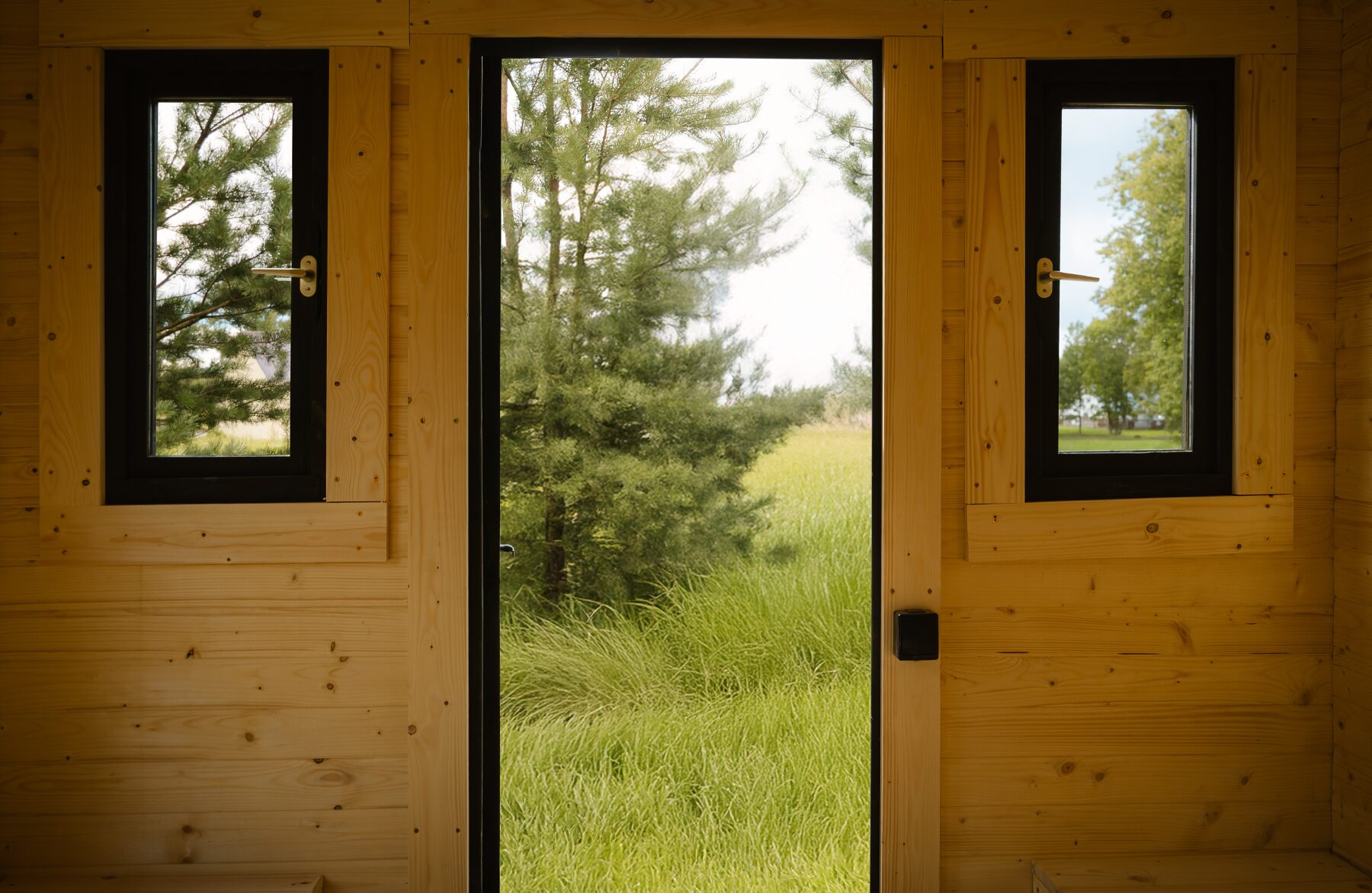 Innenansicht eines Feuerbad Tiny House CERES mit zentraler Eingangstür und zwei schmalen Fenstern auf beiden Seiten. Die helle Holzverkleidung sorgt für eine warme, natürliche Atmosphäre, während der Blick durch Tür und Fenster auf Wiese und Bäume eine starke Verbindung zur umgebenden Natur schafft