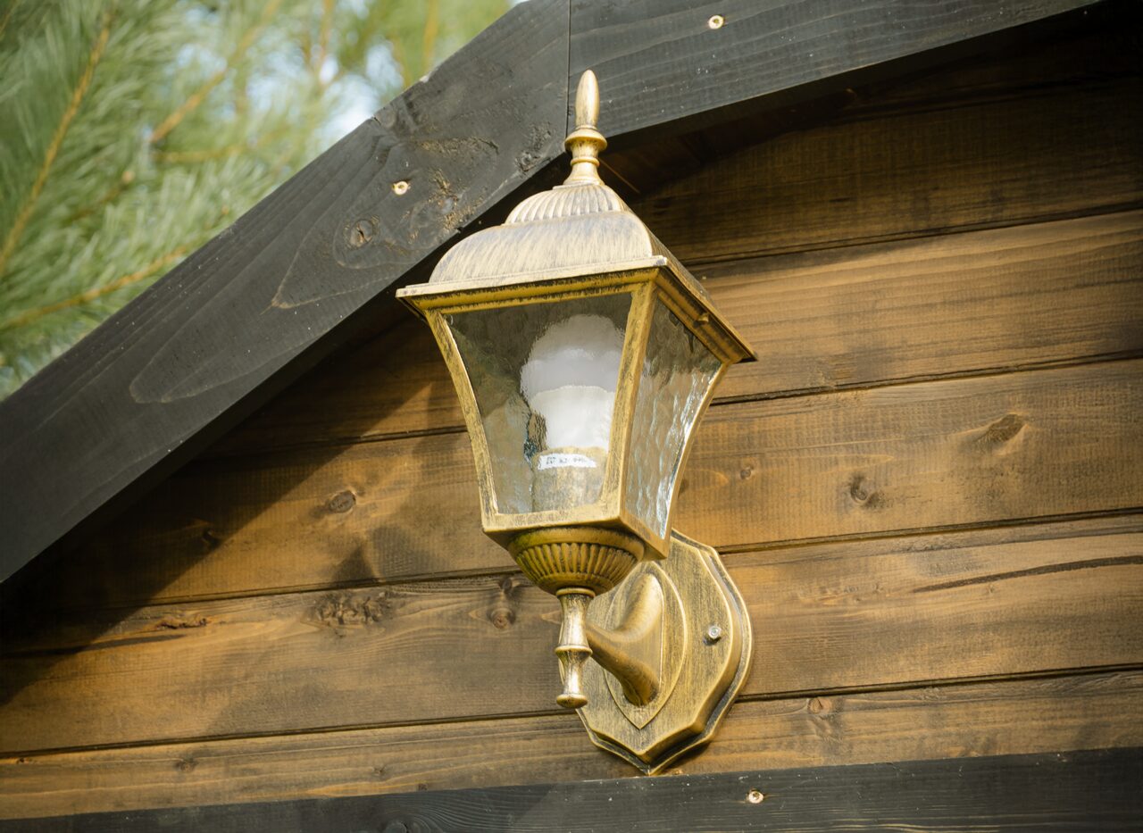 Detailaufnahme einer klassischen Außenwandleuchte aus Metall und Glas, montiert an der Holzfassade eines Feuerbad Tiny House CERES. Die Leuchte ist oberhalb des Eingangs angebracht und ergänzt die rustikale Optik der dunklen Holzverkleidung im Außenbereich