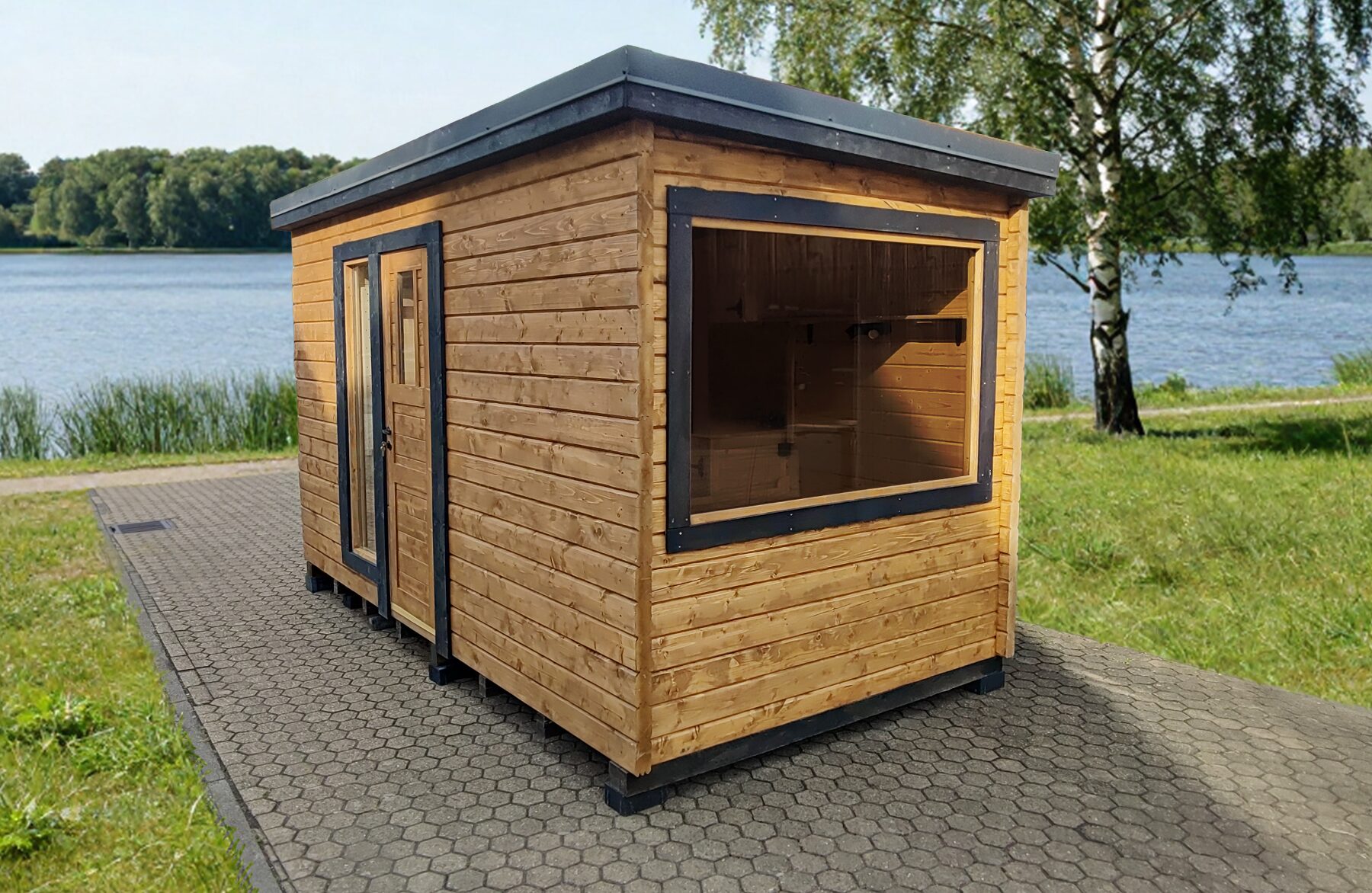 Außenansicht eines Feuerbad Tiny House DELUXE mit Holzverkleidung und dunklen Fensterrahmen, auf gepflastertem Untergrund direkt am See, umgeben von grüner Wiese, Bäumen und ruhiger Naturlandschaft
