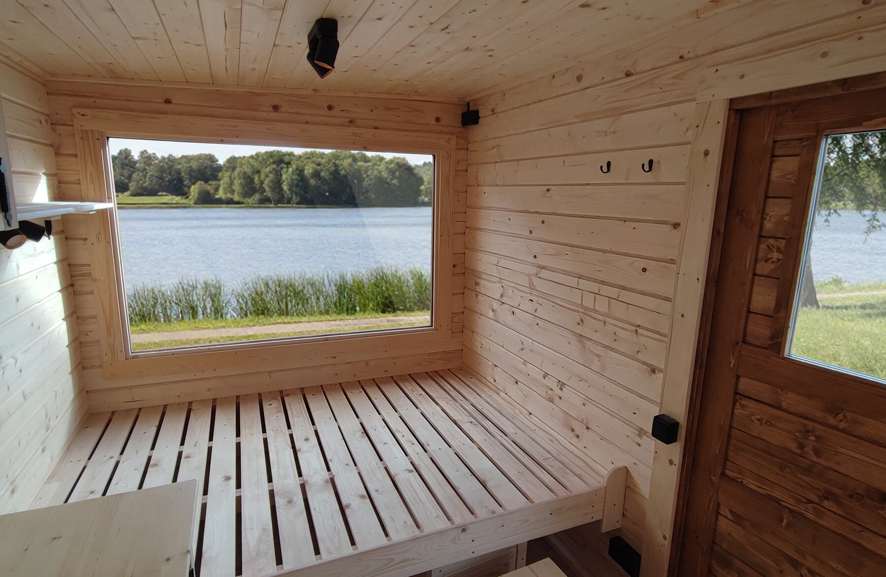 Innenansicht des Feuerbad Tiny House DELUXE mit hellem Holzinterieur, großzügiger Liegefläche und großem Panoramafenster mit Blick auf einen See und Schilf, lichtdurchflutet und naturnah gestaltet