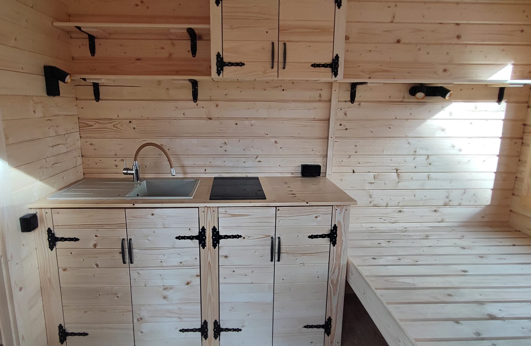 Innenansicht eines Feuerbad Tiny House DELUXE mit kompakter Holzküche, bestehend aus Spüle, Arbeitsfläche mit Kochfeld, Unterschränken und offenen Regalen. Die helle Holzverkleidung, schwarzen Beschläge und integrierten Leuchten schaffen eine funktionale und gemütliche Wohnatmosphäre