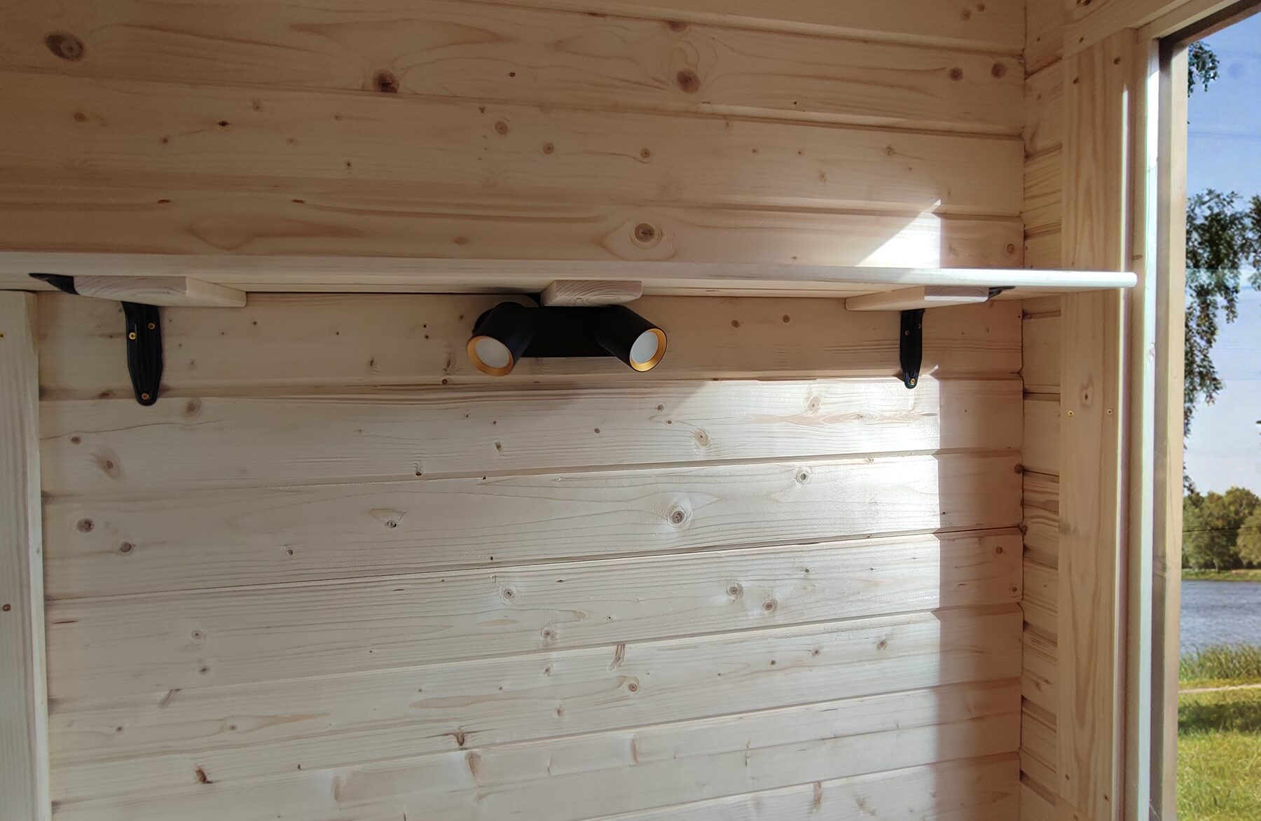 Detailansicht der Innenwand eines Feuerbad Tiny House DELUXE mit heller Holzverkleidung, schmalem Wandregal auf schwarzen Metallhalterungen und integrierter schwarzer Doppel-Wandleuchte. Seitlich fällt Tageslicht durch ein Fenster und unterstreicht die natürliche Wohnatmosphäre