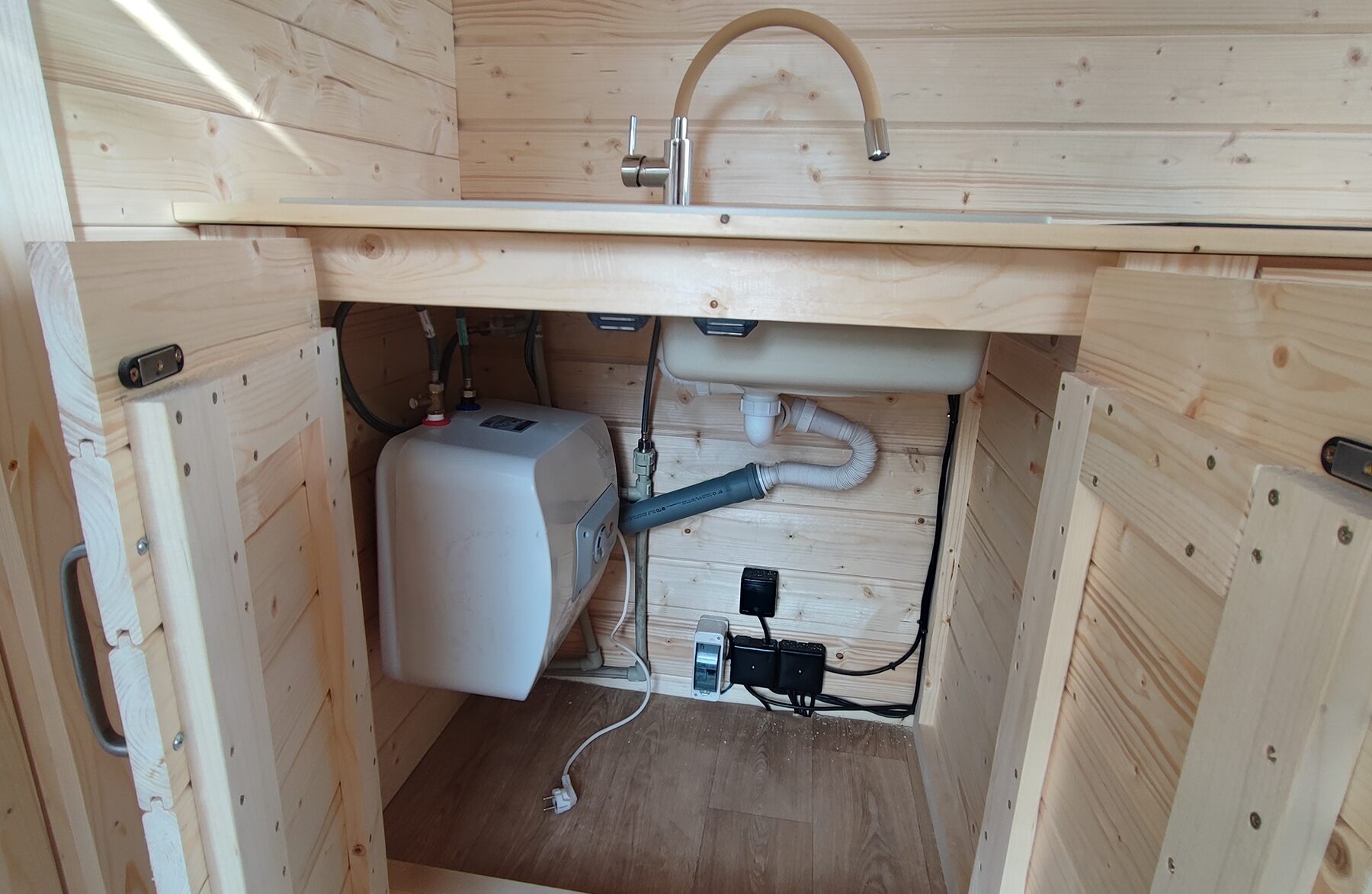 Detailansicht des Küchenbereichs in einem Feuerbad Tiny House DELUXE mit geöffneten Unterschranktüren: Zu sehen sind das Spülbecken mit Armatur, die Wasserinstallation inklusive Warmwasserboiler, Abflussleitungen sowie elektrische Anschlüsse. Die helle Holzverkleidung unterstreicht die funktionale und platzsparende Bauweise