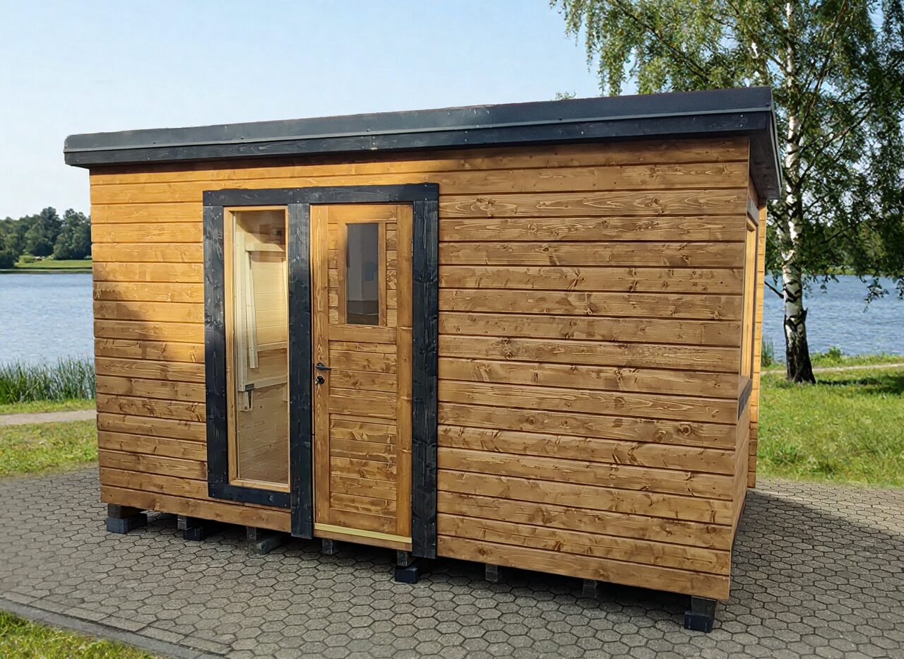Außenansicht eines Feuerbad Tiny House DELUXE mit Holzverkleidung und dunklen Fensterrahmen, auf gepflastertem Untergrund direkt am See, umgeben von grüner Wiese, Bäumen und ruhiger Naturlandschaft