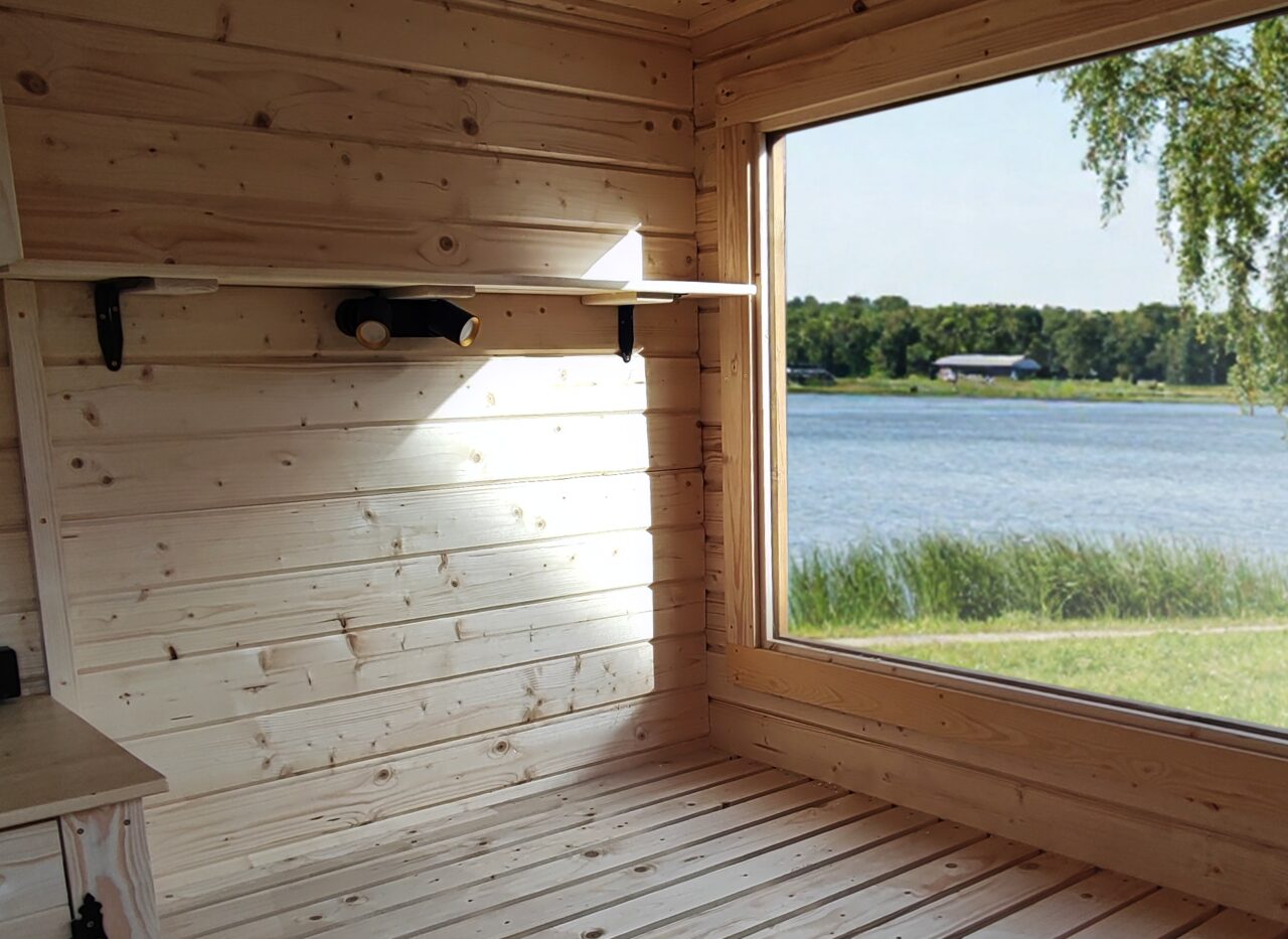 Innenansicht des Feuerbad Tiny House DELUXE mit hellem Holzinterieur, großzügiger Liegefläche und großem Panoramafenster mit Blick auf einen See und Schilf, lichtdurchflutet und naturnah gestaltet