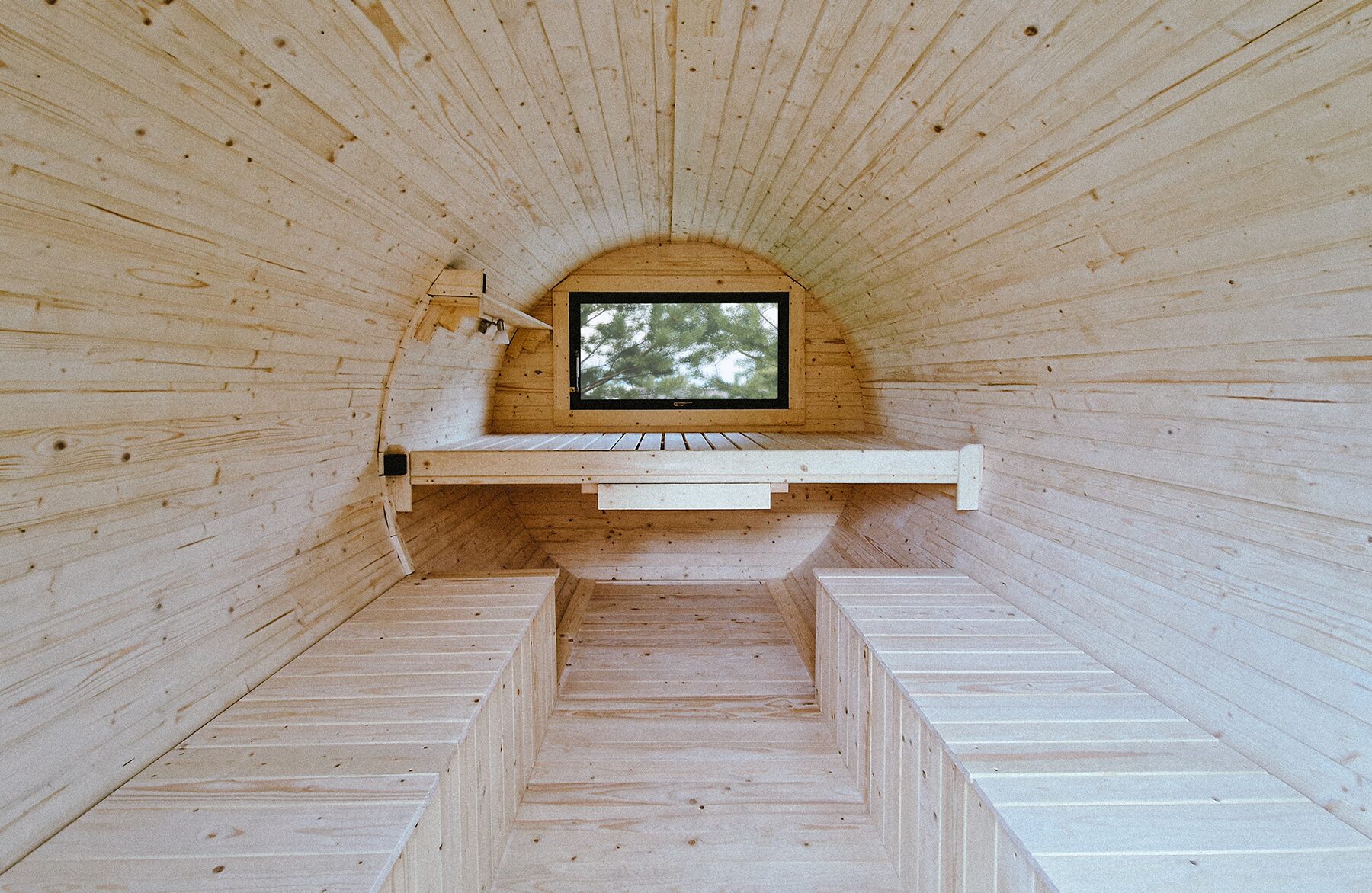 Innenansicht des Feuerbad Tiny House JUNO mit heller Holzverkleidung, zwei gegenüberliegenden Sitzbänken und einer erhöhten Liegefläche am Ende. Ein kleines Fenster sorgt für Tageslicht und Blick ins Grüne
