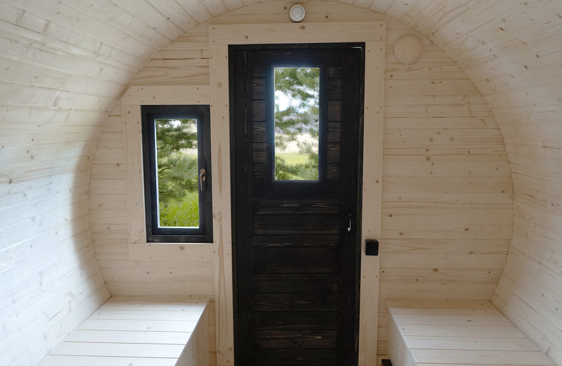 Innenansicht des Feuerbad Tiny House JUNO mit heller Holzverkleidung, schwarzer Eingangstür mit schmalem Sichtfenster und einem seitlichen Fenster. Zwei helle Sitzbänke links und rechts bieten Platz zum Entspannen, während Tageslicht und Naturblick für eine ruhige Atmosphäre sorgen