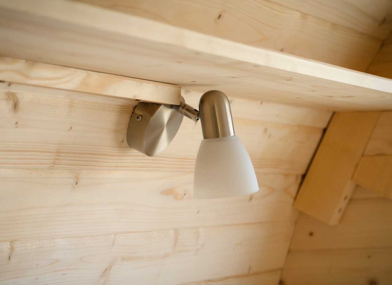 Detailaufnahme der Innenbeleuchtung des Feuerbad Tiny House JUNO. Eine moderne, schwenkbare Wandleuchte aus Metall mit mattem Glasschirm ist an der hellen Holzverkleidung montiert und sorgt für warmes, indirektes Licht im Innenraum
