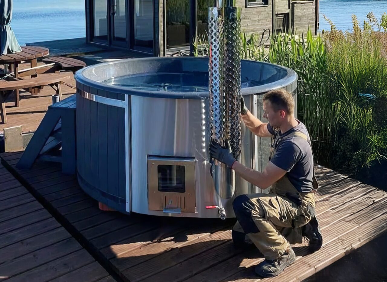 Ein Monteur installiert einen Edelstahlofen an einem Hot Tub von Feuerbad auf einer Holzterrasse direkt am Wasser