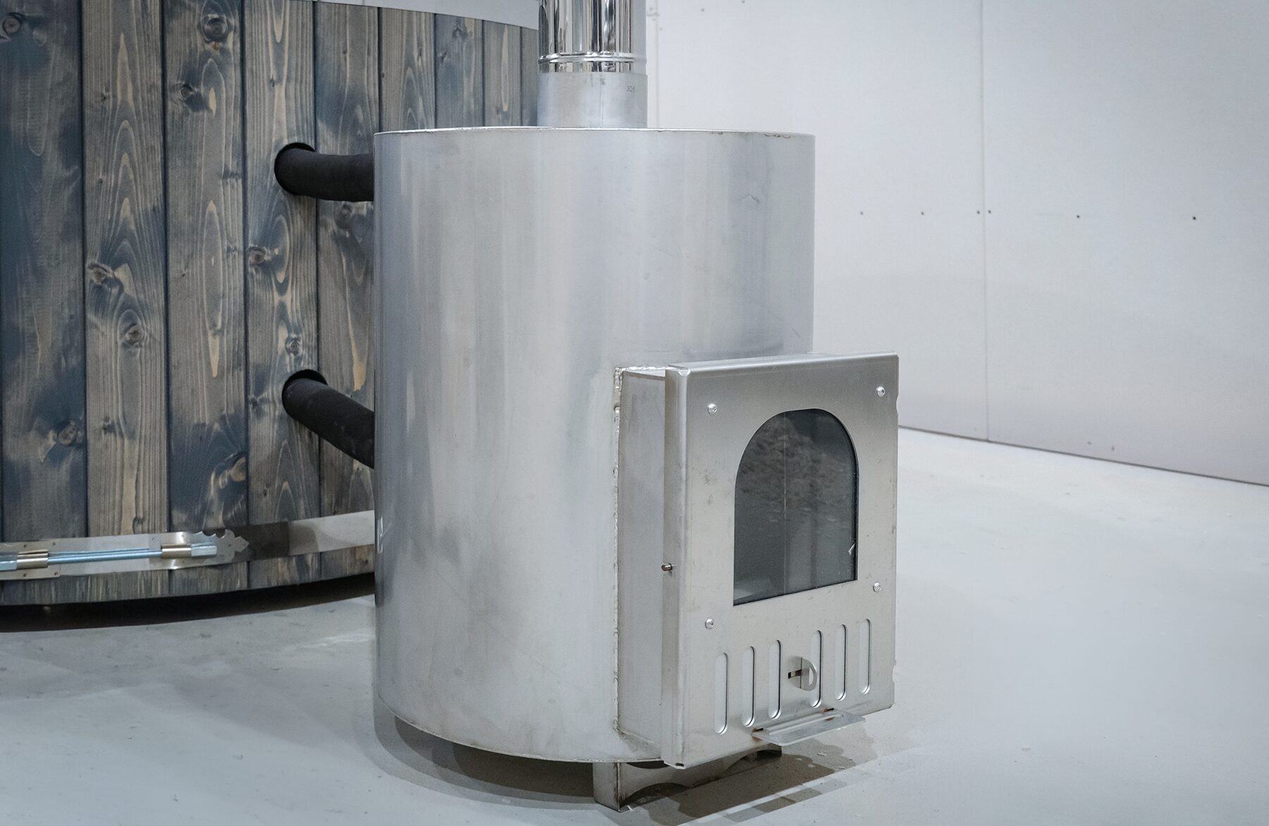 Detailansicht des externen Metallofens des Feuerbad Hot Tubs DIANA, mit Feuerraumtür, Anschlussleitungen zum Badefass und robuster Konstruktion neben der Holzverkleidung.