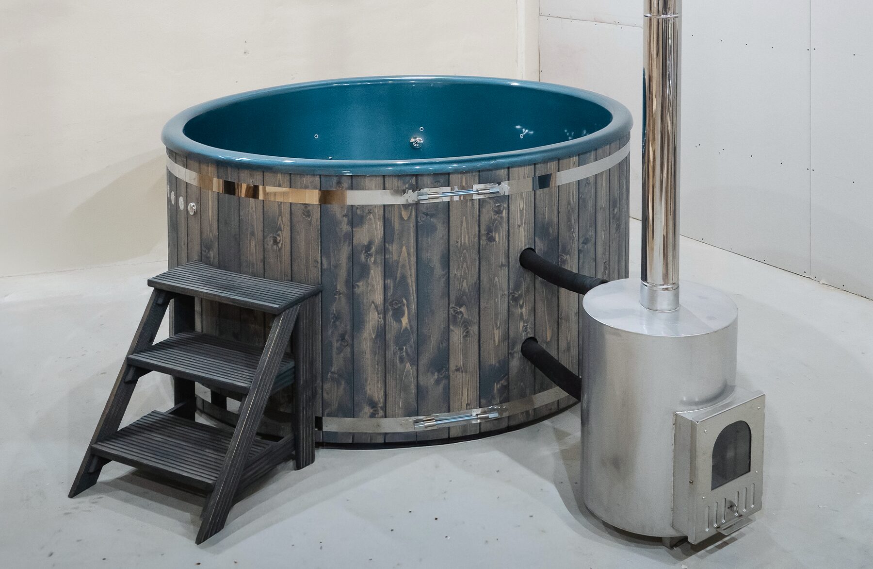 Feuerbad Hot Tub DIANA steht in einer Raumecke und verfügt über eine Holzverkleidung, eine Holz-Einstiegstreppe, eine isolierende Abdeckung sowie einen externen Metallofen mit hohem Schornstein.