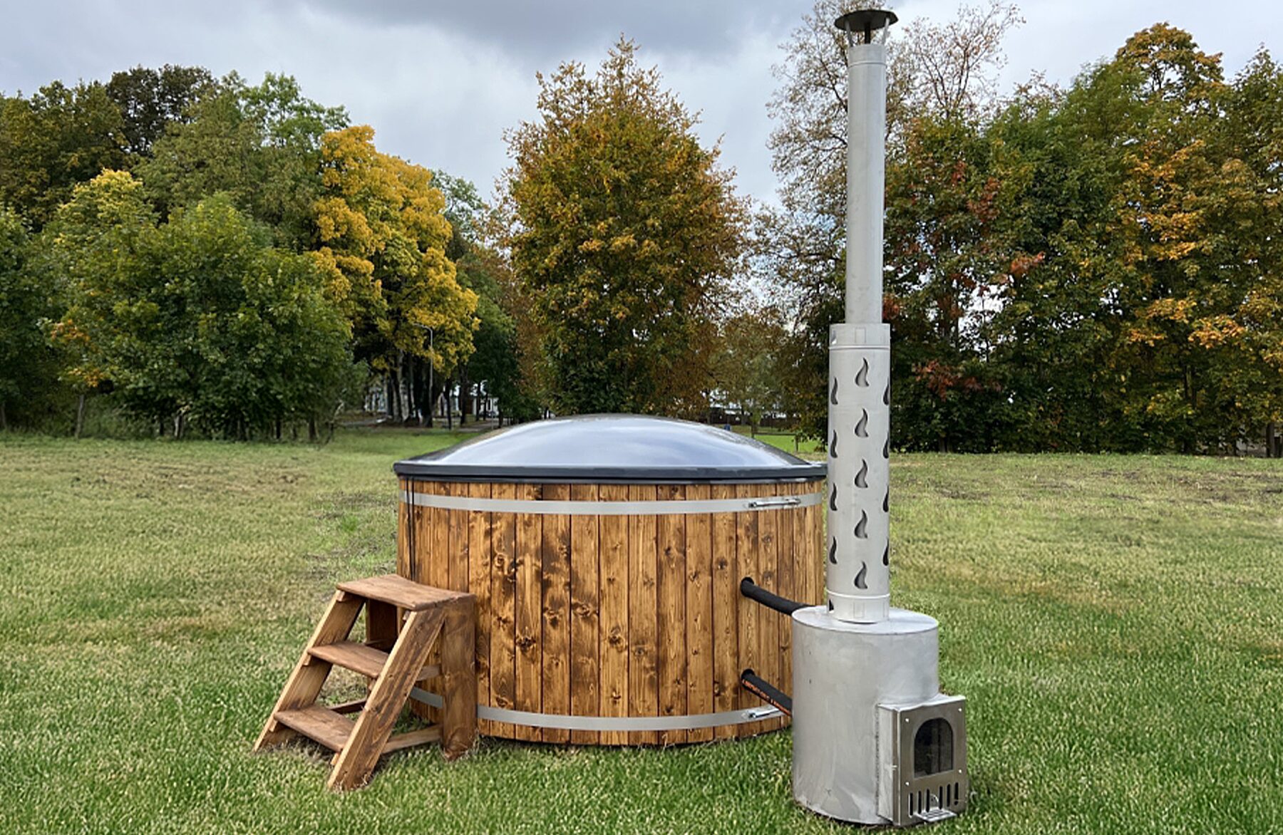 Feuerbad Hot Tub DIANA steht auf einer grünen Wiese und verfügt über eine Holzverkleidung, eine Holz-Einstiegstreppe, eine isolierende Abdeckung sowie einen externen Metallofen mit hohem Schornstein, umgeben von einer natürlichen Landschaft.