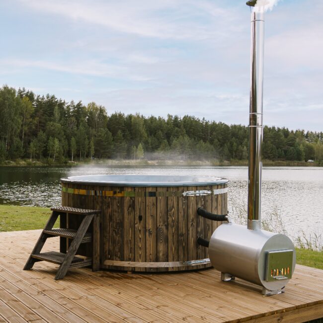 Feuerbad Hot Tub LUNA aus Holz auf einem Holzdeck, mit externem Metallofen und Schornstein, Holz-Einstiegstreppe und grüner Naturlandschaft im Hintergrund. Zwei Personen entspannen sich im dampfenden Hot Tub.